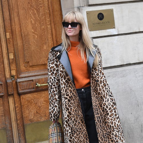 A blonde woman in an orange sweater, black pants, and a long leopard animal print coat 