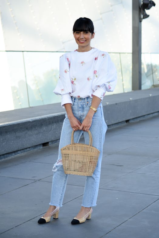 A woman in a white floral top and light denim jeans, holding a straw bag while posing for a photo